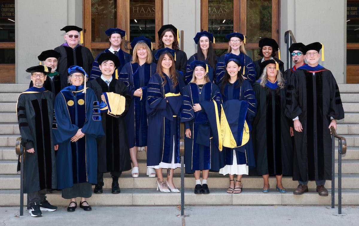 Smiling people standing outside on steps in front of a building, all wearing graduation caps and gowns.
